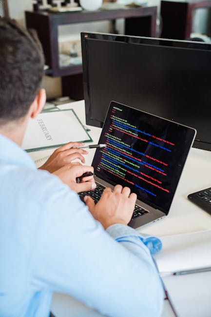 A person seated at a desk is actively engaged in computer coding or software development, with their left hand on a keyboard and their right hand holding a stylus or pointing device. The individual ha